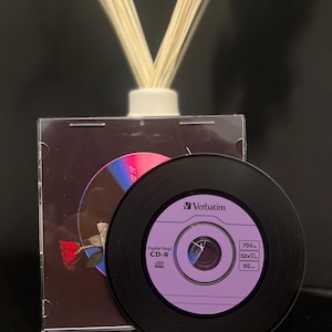 Op de afbeelding: Een zwarte CD-R-schijf met een paars label en het Verbatim-logo, naast een CD-hoesje met een rozenafbeelding. De CD-R heeft de tekst "Digital Vinyl CD-R" en afmetingen van 700 MB, 52x en 80.