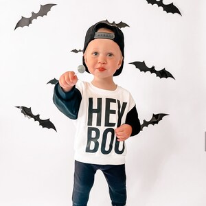 May include: A child wearing a black baseball cap, a white and black "HEY BOO" sweatshirt, black pants, and white sneakers. The child is holding a lollipop. Black bat decorations are on the white background.