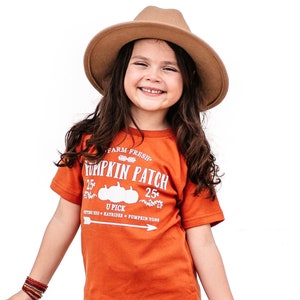 May include: A young person wearing an orange t-shirt with "Pumpkin Patch" text, a brown hat, distressed blue jeans, and brown boots. The shirt has a graphic of pumpkins. A scrunchie and bracelets complete the outfit.