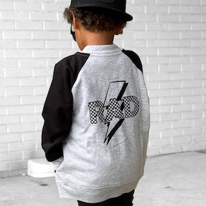 Op de afbeelding: Een kind draagt een zwarte baseballpet, een zwarte broek en witte sneakers. De grijze en zwarte sweatshirt heeft het woord "RAD" in een geblokt patroon met een bliksemgrafiek op de rug.