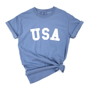 以下が含まれることがあります： 前面に白い文字で「USA」がプリントされた青いTシャツです。