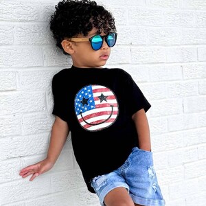 Op de afbeelding: Een zwart t-shirt met een smiley-gezicht ontwerp met de Amerikaanse vlag. Het kind draagt een blauwe zonnebril, een spijkerbroek, witte sneakers en gestreepte sokken. Het kind leunt tegen een witte bakstenen muur.
