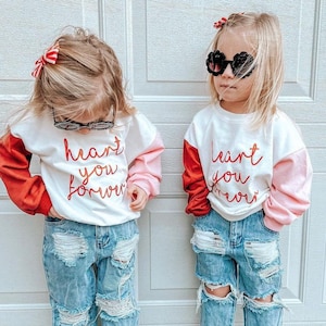 May include: Two young children wearing matching outfits. Each child is wearing a white sweatshirt with red sleeves and pink accents, and the words "heart you forever" in red script. They are also wearing distressed blue jeans and white sneakers. One child is wearing sunglasses.