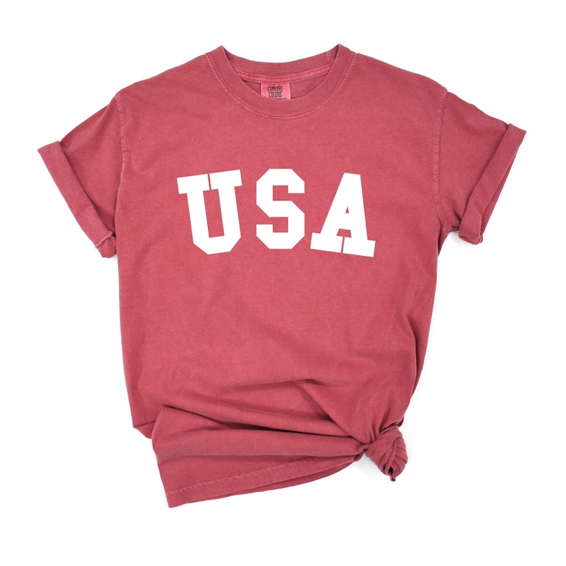 以下が含まれることがあります： 前面に白い「USA」の文字がプリントされた赤いTシャツ。柔らかく快適な素材で作られており、ゆったりとしたフィット感です。