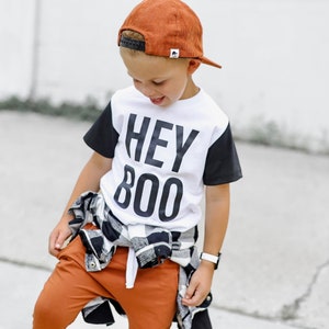 May include: A child wearing a white t-shirt with black sleeves and the text "HEY BOO" in large black letters. The child is also wearing a rust-colored baseball cap, rust-colored pants, and a black and white plaid shirt tied around the waist.