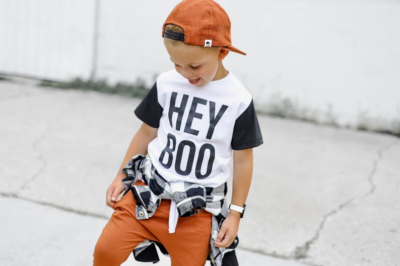 May include: A child wearing a white t-shirt with black sleeves and the text "HEY BOO" in large black letters. The child is also wearing a rust-colored baseball cap, rust-colored pants, and a black and white plaid shirt tied around the waist.