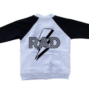Op de afbeelding: Een grijs raglan-sweatshirt met zwarte mouwen. De achterkant toont het woord "RAD" in een geblokt patroon, over een zwarte bliksem. De sweatshirt is gemaakt van een zacht, comfortabel materiaal.