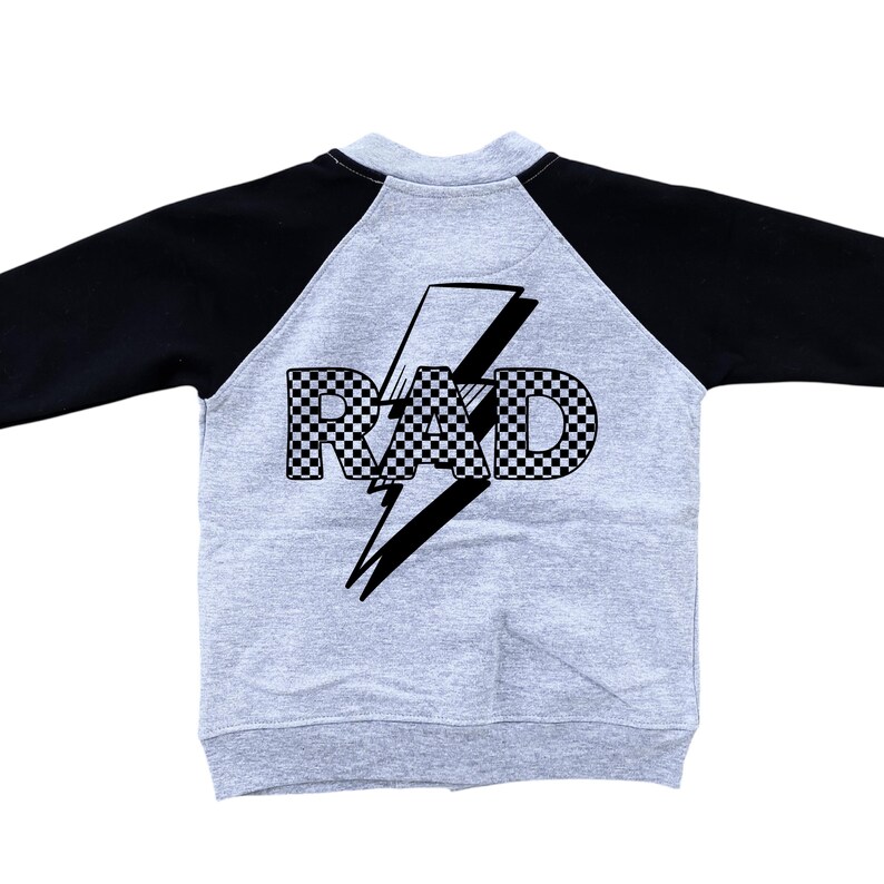Op de afbeelding: Een grijs raglan-sweatshirt met zwarte mouwen. De achterkant toont het woord "RAD" in een geblokt patroon, over een zwarte bliksem. De sweatshirt is gemaakt van een zacht, comfortabel materiaal.