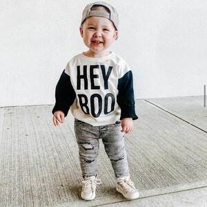 May include: A smiling toddler wearing a gray baseball cap, a white and black "HEY BOO" sweatshirt, distressed gray jeans, and beige sneakers. The child is standing on a concrete surface.