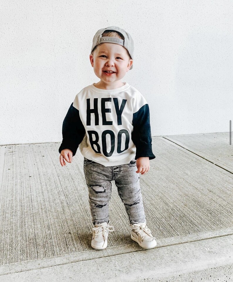 May include: A smiling toddler wearing a gray baseball cap, a white and black "HEY BOO" sweatshirt, distressed gray jeans, and beige sneakers. The child is standing on a concrete surface.