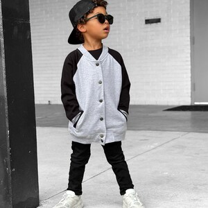 Op de afbeelding: Een kind draagt een grijs en zwart baseballjack, een zwarte broek, een zwarte pet en een zonnebril. De jas heeft een knoopsluiting en zakken. Het kind draagt ook witte sneakers.