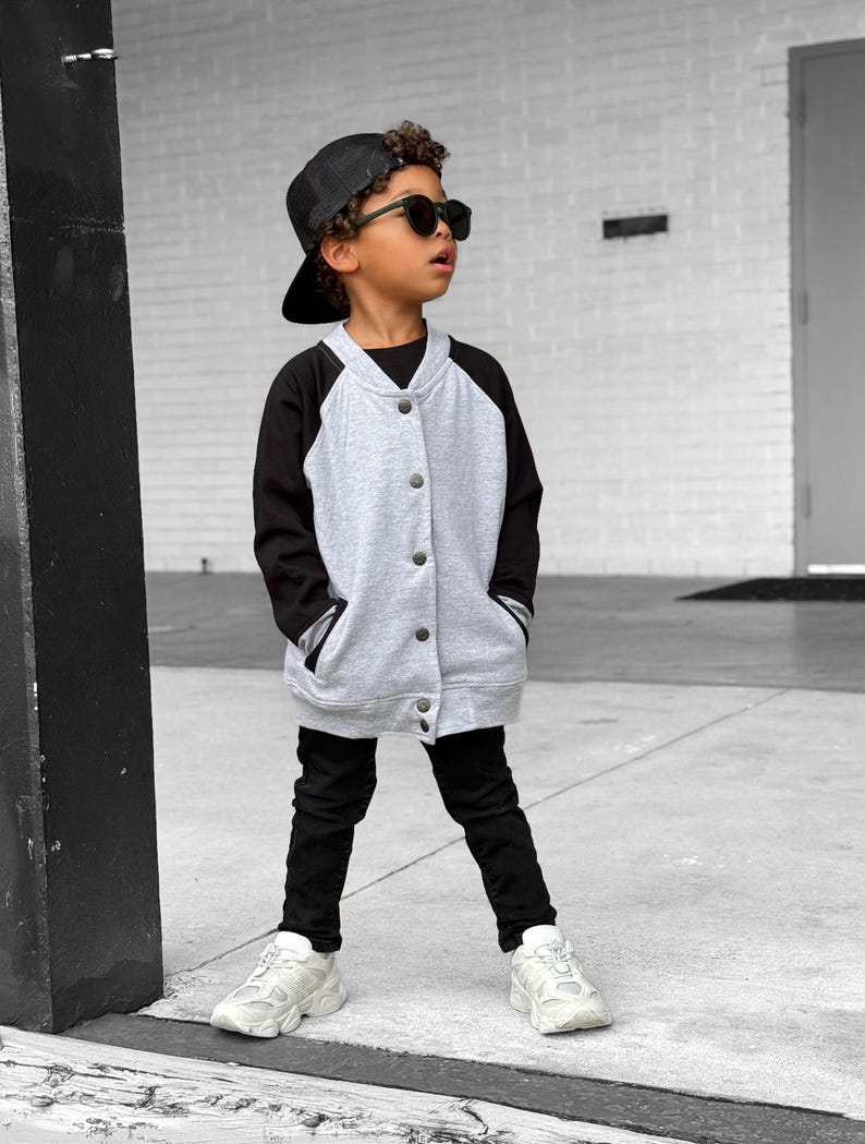 Op de afbeelding: Een kind draagt een grijs en zwart baseballjack, een zwarte broek, een zwarte pet en een zonnebril. De jas heeft een knoopsluiting en zakken. Het kind draagt ook witte sneakers.