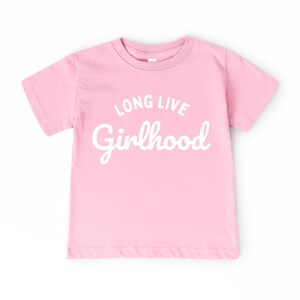 May include: A light pink t-shirt with the words "LONG LIVE Girlhood" printed in white script. The shirt has short sleeves and a crew neck. The text is centered on the front of the shirt.