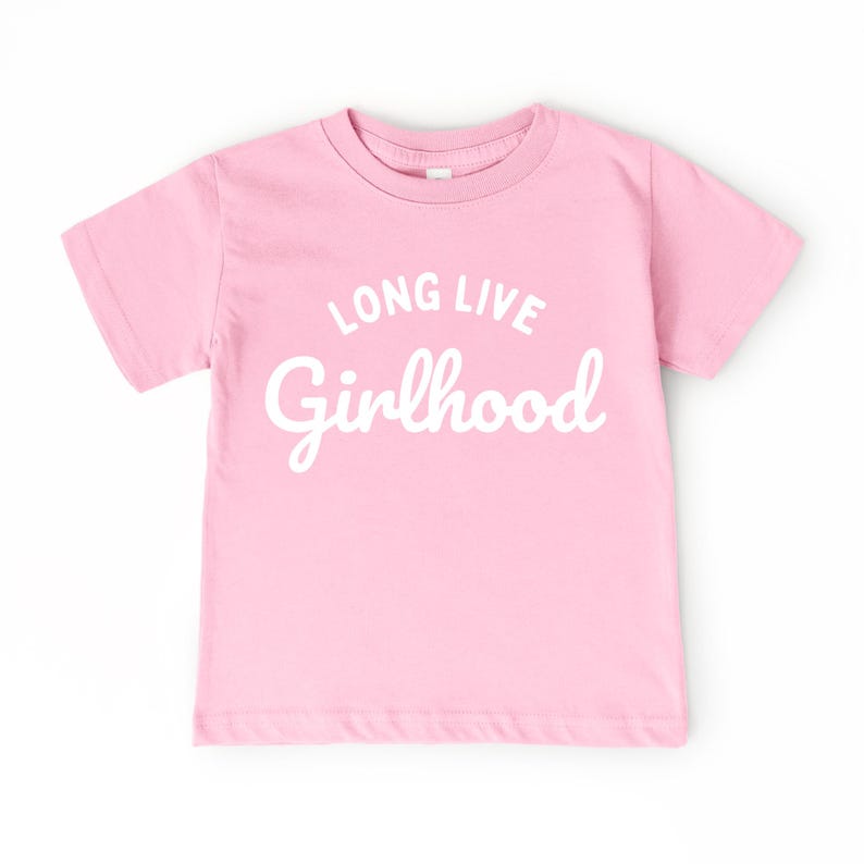 May include: A light pink t-shirt with the words "LONG LIVE Girlhood" printed in white script. The shirt has short sleeves and a crew neck. The text is centered on the front of the shirt.