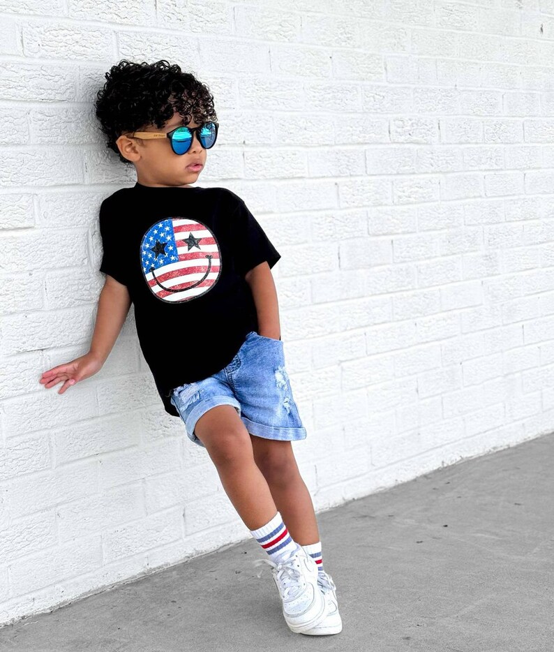 Op de afbeelding: Een zwart t-shirt met een smiley-gezicht ontwerp met de Amerikaanse vlag. Het kind draagt een blauwe zonnebril, een spijkerbroek, witte sneakers en gestreepte sokken. Het kind leunt tegen een witte bakstenen muur.