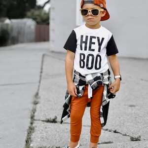May include: A child wearing a burnt orange baseball cap, sunglasses, and a white t-shirt with black sleeves and the words "HEY BOO". They are also wearing orange pants, a black and white plaid shirt tied around the waist, and checkered slip-on shoes.