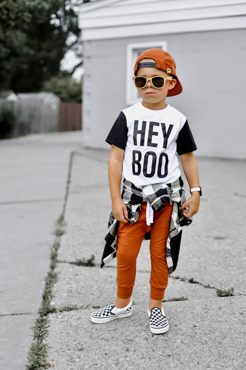 May include: A child wearing a burnt orange baseball cap, sunglasses, and a white t-shirt with black sleeves and the words "HEY BOO". They are also wearing orange pants, a black and white plaid shirt tied around the waist, and checkered slip-on shoes.