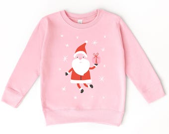 Può includere: Una felpa a maniche lunghe rosa chiaro con una grafica centrale di un Babbo Natale in stile cartoon con abito e cappello rossi, che tiene in mano un piccolo regalo rosa. Il disegno è circondato da fiocchi di neve bianchi a forma di stella.