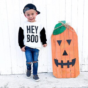 May include: A child wearing a black baseball cap, a white and black long-sleeved shirt with the text "HEY BOO", and ripped blue jeans. A large orange wooden pumpkin decoration with a jack-o'-lantern face stands nearby.
