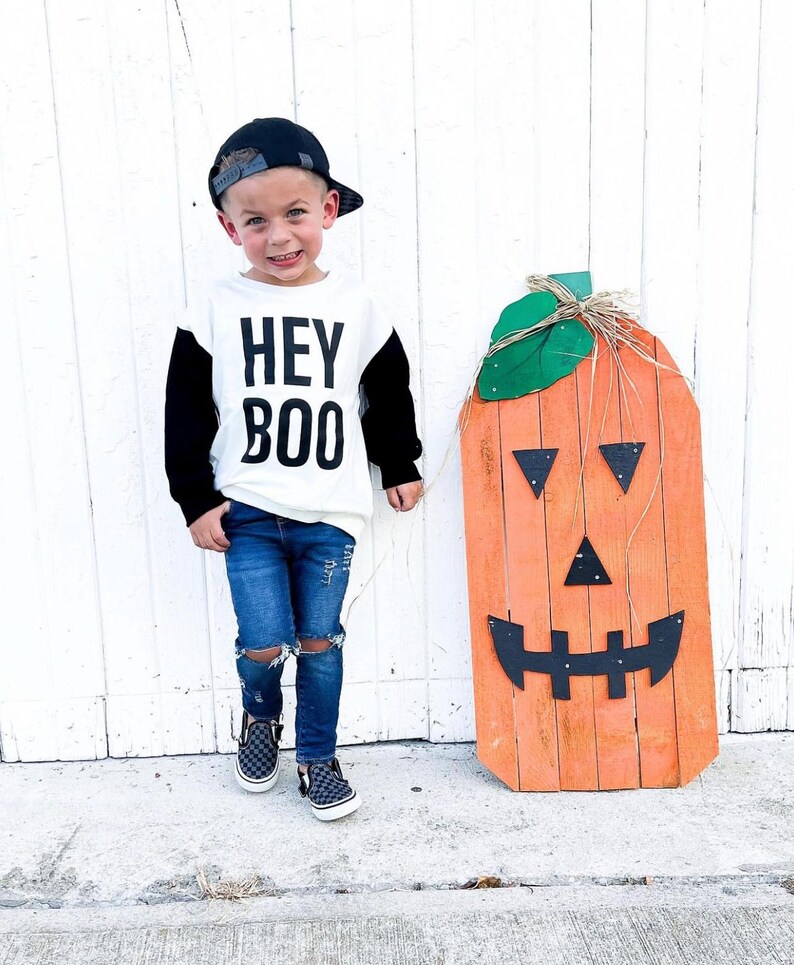 May include: A child wearing a black baseball cap, a white and black long-sleeved shirt with the text "HEY BOO", and ripped blue jeans. A large orange wooden pumpkin decoration with a jack-o'-lantern face stands nearby.