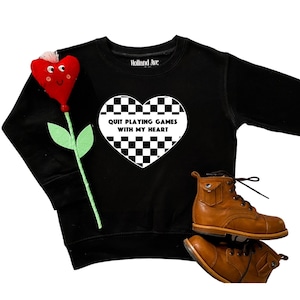 May include: Black sweatshirt with a white checkered heart graphic that reads "QUIT PLAYING GAMES WITH MY HEART." A red felt flower and brown leather boots are also in the image. The sweatshirt is from Holland Ave.