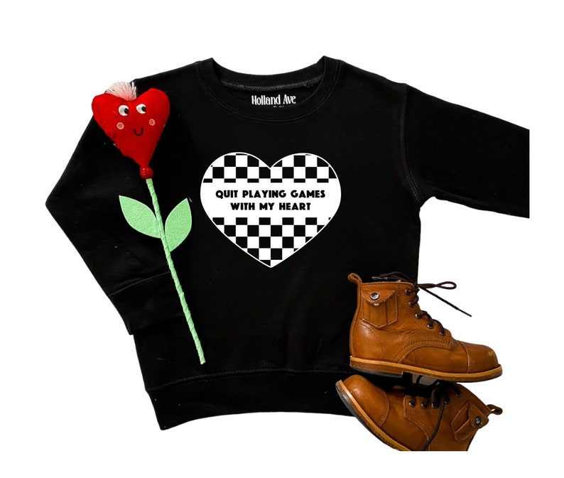 May include: Black sweatshirt with a white checkered heart graphic that reads "QUIT PLAYING GAMES WITH MY HEART." A red felt flower and brown leather boots are also in the image. The sweatshirt is from Holland Ave.