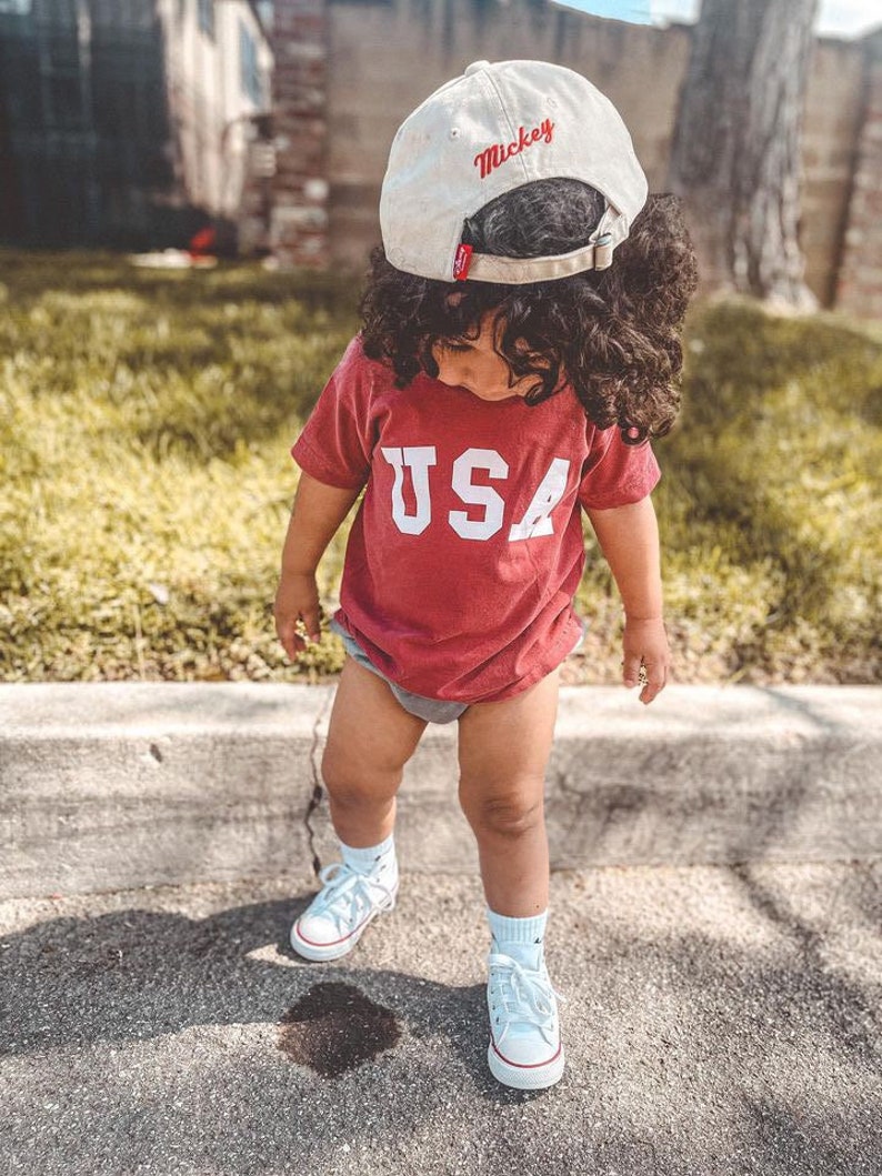 以下が含まれることがあります： 赤いシャツを着て、白文字で「USA」と書かれた小さな子供。子供は赤い文字で「ミッキー」と書かれた白い野球帽もかぶっています。子供は白いスニーカーを履いていて、歩道に立っています。