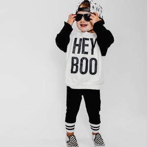 May include: A child's outfit featuring a white sweatshirt with black sleeves and the text "HEY BOO" in bold black letters. The outfit includes black pants with white stripes at the ankles, a baseball cap with lightning bolt pattern, and checkered sneakers. The child is wearing sunglasses.