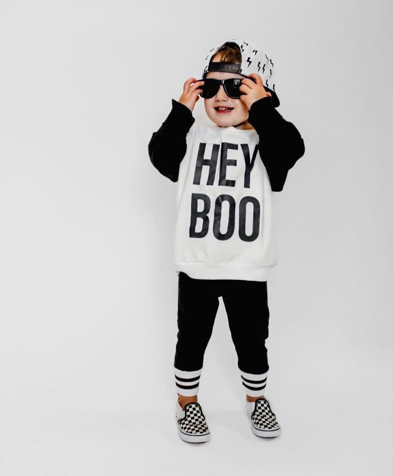 May include: A child's outfit featuring a white sweatshirt with black sleeves and the text "HEY BOO" in bold black letters. The outfit includes black pants with white stripes at the ankles, a baseball cap with lightning bolt pattern, and checkered sneakers. The child is wearing sunglasses.