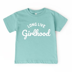 May include: A light teal short-sleeved t-shirt with the words "LONG LIVE Girlhood" printed in white. The shirt has a crew neck and is made of a soft material.