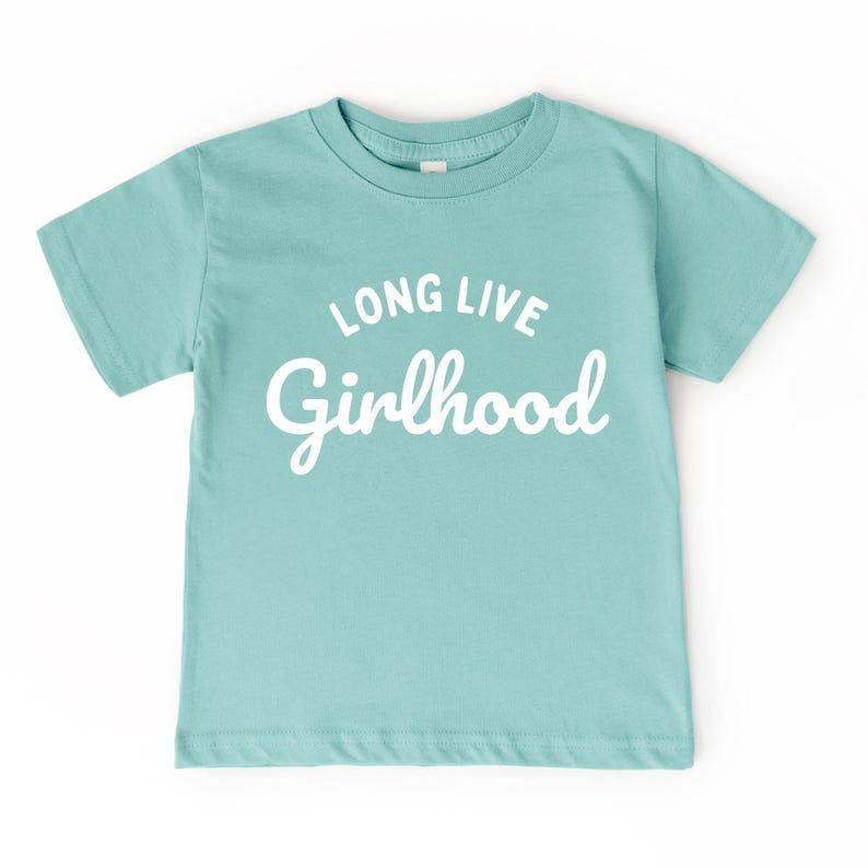 May include: A light teal short-sleeved t-shirt with the words "LONG LIVE Girlhood" printed in white. The shirt has a crew neck and is made of a soft material.