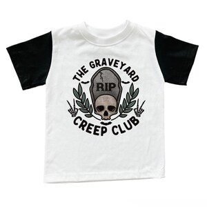 Puede incluir: Una camiseta blanca con mangas negras que presenta un gráfico de una lápida con el texto RIP, una calavera, dos ramas con hojas y manos de esqueleto haciendo el signo de la paz. El texto THE GRAVEYARD CREEP CLUB está impreso en un arco circular.