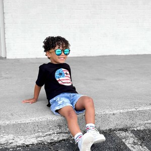 Op de afbeelding: Een jong persoon draagt een zwart t-shirt met een smiley-gezichtontwerp met de Amerikaanse vlag, gecombineerd met een spijkerbroek en witte sneakers. De persoon draagt ook een zonnebril met een houten montuur.