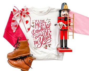Può includere: Un completo a tema festivo con una felpa bianca con la scritta "It's the Most Wonderful Time of the Year". L'outfit include un fiocco rosso, stivali marroni e uno schiaccianoci rosso e nero. La felpa ha maniche lunghe rosa.