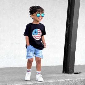 Op de afbeelding: Een kind draagt een zwart t-shirt met een smiley-ontwerp van de Amerikaanse vlag, een blauwe spijkerbroek, witte sneakers en een zonnebril. De outfit wordt gecompleteerd met witte sokken met rode en blauwe strepen.