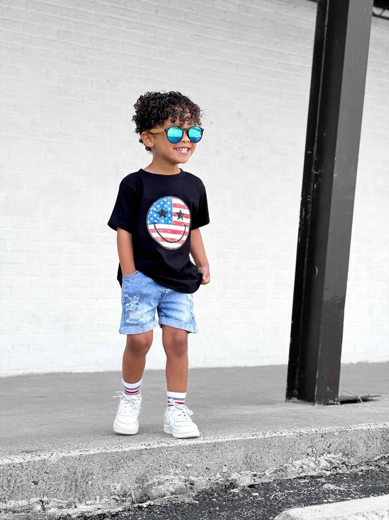 Op de afbeelding: Een kind draagt een zwart t-shirt met een smiley-ontwerp van de Amerikaanse vlag, een blauwe spijkerbroek, witte sneakers en een zonnebril. De outfit wordt gecompleteerd met witte sokken met rode en blauwe strepen.