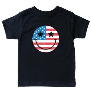 Op de afbeelding: Een marineblauw T-shirt met een versleten smiley-gezicht ontwerp. De smiley bevat de Amerikaanse vlag, met sterren en strepen, en twee zwarte sterrenogen. Het ontwerp is gecentreerd op de voorkant van het shirt.