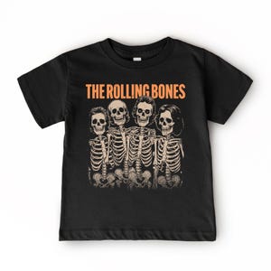 Puede incluir: Una camiseta negra de manga corta con un estampado gráfico de cuatro esqueletos con pelo, dispuestos en fila. Sobre los esqueletos, el texto THE ROLLING BONES está impreso en una fuente naranja, negrita y sin remates.