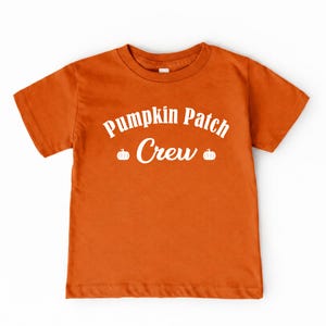 May include: A short-sleeved orange t-shirt featuring the white text Pumpkin Patch Crew printed across the chest. The design includes two small white pumpkin icons, one on each side of the word Crew, centered on the front of the garment.