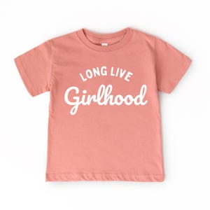 May include: A short-sleeved, coral-colored t-shirt with the words "LONG LIVE Girlhood" in white script. The shirt is made of a soft material and is suitable for children.