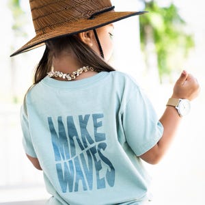 May include: A light blue t-shirt with the text "MAKE WAVES" printed in a blue gradient on the back. The shirt is being worn by a child wearing a brown straw hat and blue jeans.