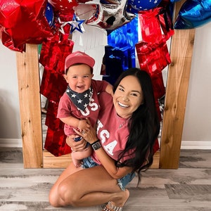 以下が含まれることがあります： 女性と小さな子供が、白い「USA」の文字が入った赤いシャツを合わせて着ています。子供は青と白の星柄のバンドゥを着用しています。女性は笑顔で子供を抱っこしています。彼らは赤、白、青の風船アーチの前に立っています。