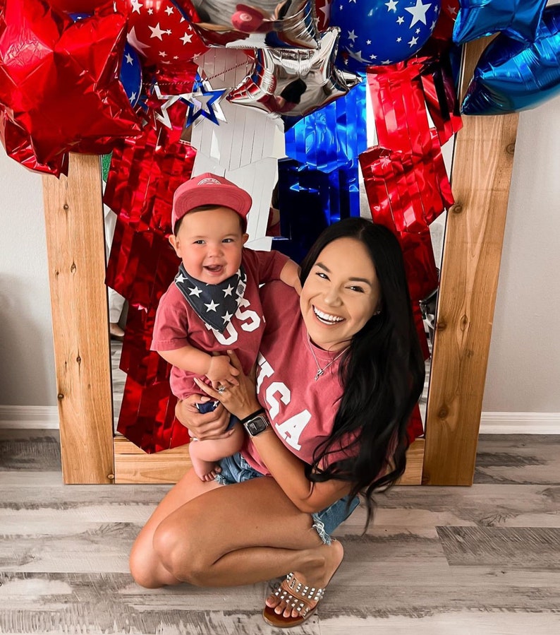 以下が含まれることがあります： 女性と小さな子供が、白い「USA」の文字が入った赤いシャツを合わせて着ています。子供は青と白の星柄のバンドゥを着用しています。女性は笑顔で子供を抱っこしています。彼らは赤、白、青の風船アーチの前に立っています。