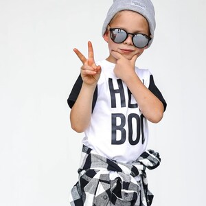 May include: A child wearing a gray beanie, round sunglasses, and a white t-shirt with the words "HEY BOO" in black. A black and white plaid shirt is tied around the waist.