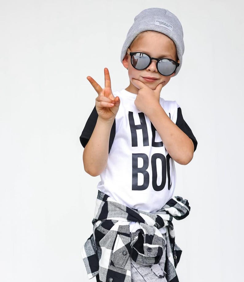 May include: A child wearing a gray beanie, round sunglasses, and a white t-shirt with the words "HEY BOO" in black. A black and white plaid shirt is tied around the waist.