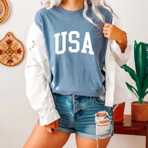 Pu&ograve; includere: Una maglietta blu con la scritta "USA" stampata in bianco sul davanti. La maglietta &egrave; indossata da una persona con una giacca bianca e pantaloncini di jeans.