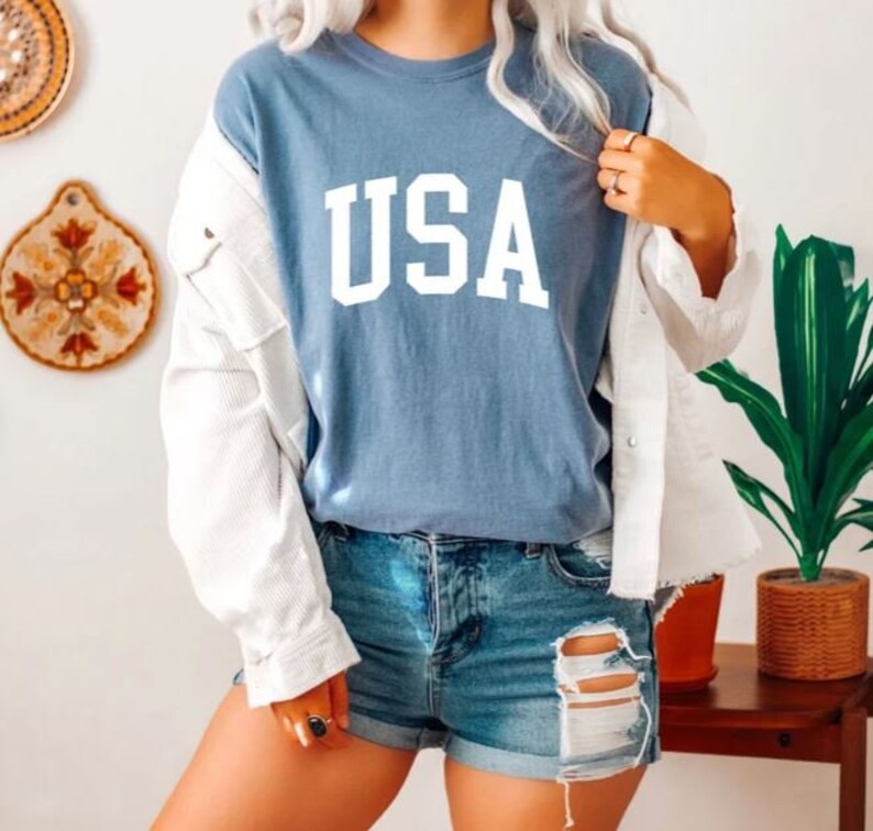 Pu&ograve; includere: Una maglietta blu con la scritta "USA" stampata in bianco sul davanti. La maglietta &egrave; indossata da una persona con una giacca bianca e pantaloncini di jeans.