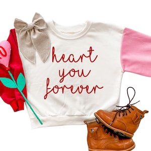 May include: A white and pink long-sleeved sweatshirt with the text "heart you forever" in red script. A tan bow and a red felt heart-shaped decoration with "XO" are next to the shirt. Brown leather boots are at the bottom.