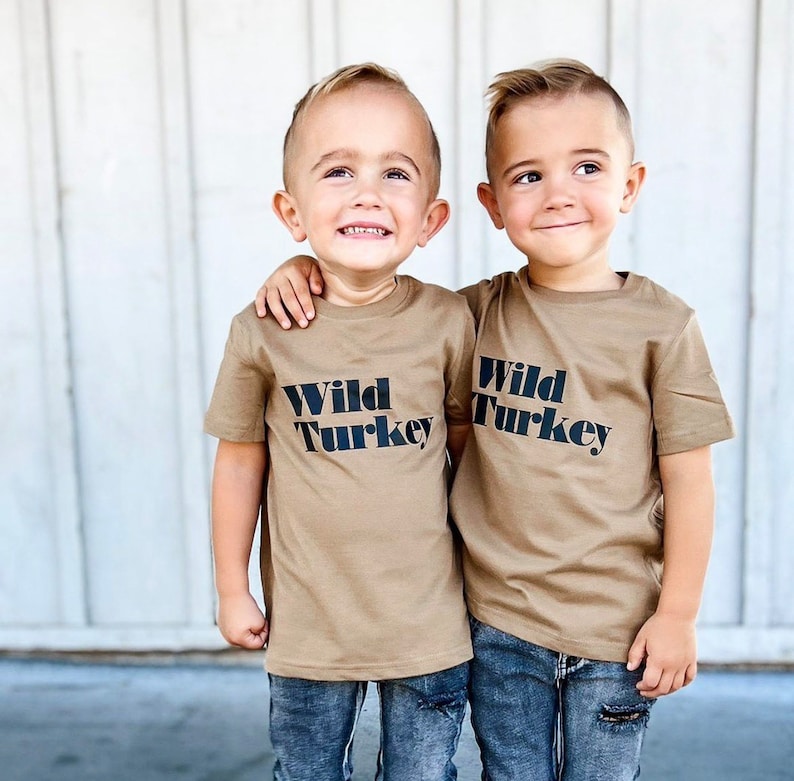 May include: Two young children wearing tan t-shirts with the text "Wild Turkey" in black. They are wearing blue jeans. One child has their arm around the other. The background is a white wooden wall.