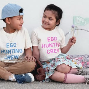 May include: Two children wearing matching cream-colored t-shirts with the text "EGG HUNTING CREW" in blue and pink. One child wears a blue baseball cap and brown pants. The other child wears a floral skirt and pink socks.
